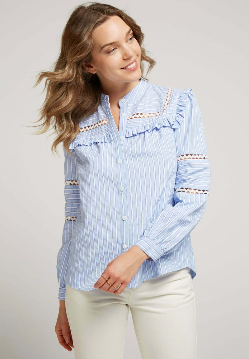 Blouse rayée bleu et blanc avec détail volanté, accents en dentelle, manches longues et fermeture à boutons. Confectionnée en tissu léger.