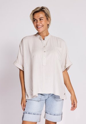 Smiling woman with short blond hair wearing a loose beige short-sleeve buttoned shirt and rolled-up light denim shorts.