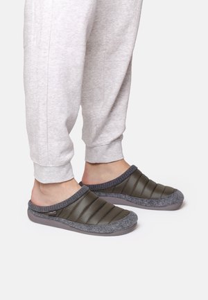 Zapatillas acolchadas verdes con borde de felpa gris y suela de goma, con un diseño de fácil acceso y textura suave, llevadas con pantalones de chándal grises.