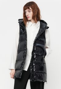 Jonge vrouw in een glanzende zwarte pufferwest met capuchon over een witte blouse en zwarte broek, kijkt opzij.