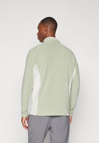 Veste en polaire vert clair avec un panneau latéral blanc, col montant et manches longues, présentant une texture lisse et un design épuré.