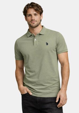 Mann mit kurzen lockigen Haaren, der ein hellgrünes Poloshirt mit dunklem Emblem trägt, Hände entspannt, vor einfachem hellem Hintergrund.