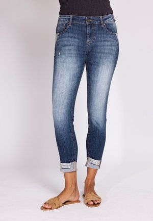 Woman wearing rolled-up, faded blue skinny jeans with slight distressing and tan open-toe flat sandals, standing against a plain background.