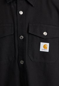 Sort skjorte med to brystlommer, sølv snapknapper og et Carhartt-logo patch med en stiliseret orange bue på en hvid baggrund.