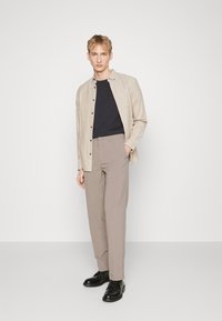 Camicia beige, t-shirt nera a girocollo e pantaloni grigi chiari. Outfit completato con scarpe nere stringate. Il modello è in piedi contro uno sfondo bianco.