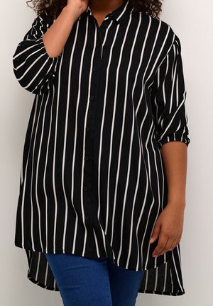 Persoon die een loszittend zwart overhemd met verticale witte strepen en een blauwe spijkerbroek draagt, staand tegen een effen lichte achtergrond.