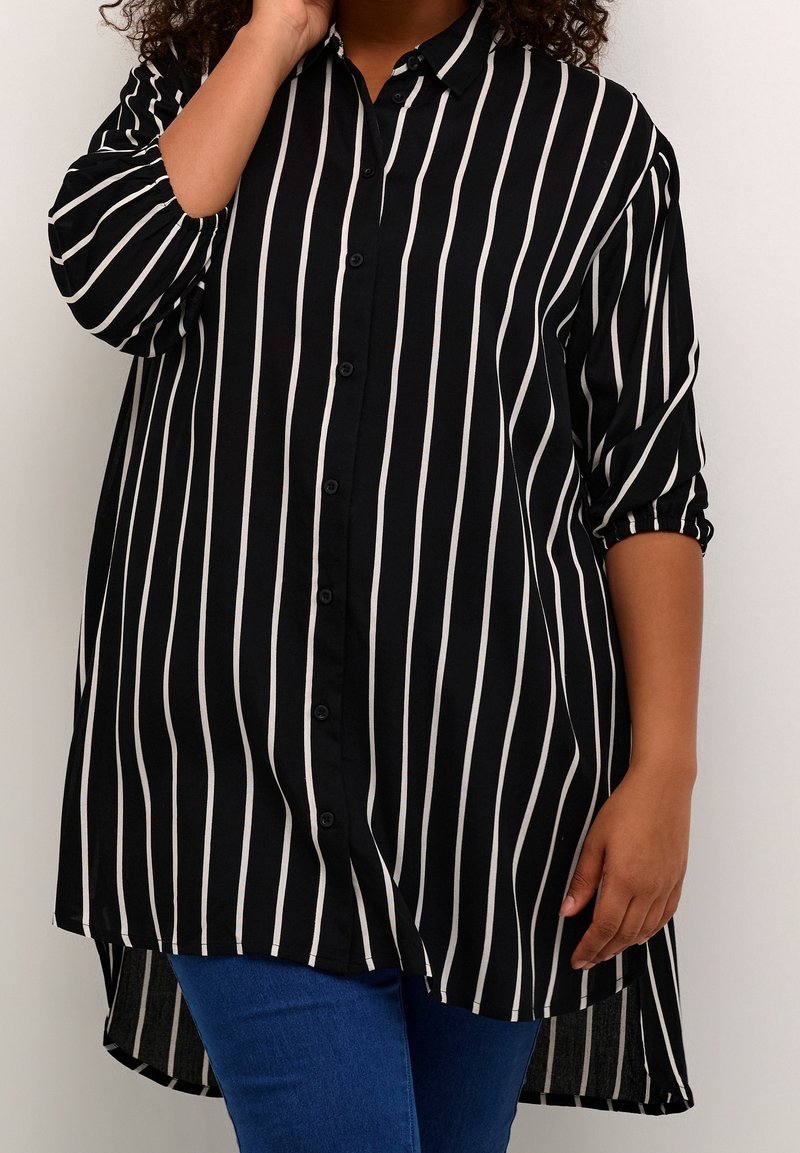 Personne portant une chemise noire ample à boutons avec des rayures blanches verticales et un jean bleu, se tenant devant un fond clair uni.