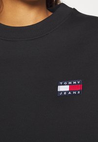 Černá mikina se žebrovaným výstřihem, která má na hrudi vyšitý modro-červený nášivku "TOMMY JEANS". Hladká textilie.