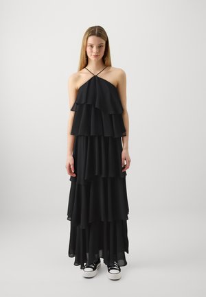 Robe noire à couches en tissu fluide, ornée de multiples volants, avec un décolleté halter, et assortie à des baskets noires.