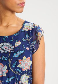 Námořnická blůza s květinovým vzorem; má krátké, průsvitné rukávy a kulatý výstřih; látka vypadá lehce a hladce.