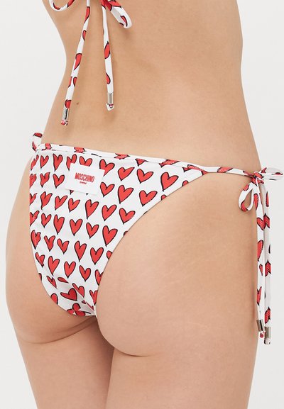 White bikini bottom with red heart pattern and side ties, featuring a Moschino Swim label on the back, worn by a person.