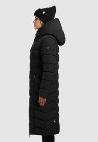 Lang zwarte pufferjas met een capuchon, voorzien van gestikte panelen, een ritssluiting aan de voorkant en een logo-patch nabij de zoom. Draagt een zwarte gebreide muts.