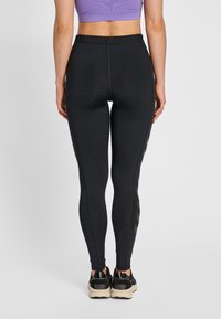 Kobieta ubrana w czarne legginsy z wysokim stanem i czarne buty sportowe, stojąca tyłem do aparatu na jednolitym jasnym tle.