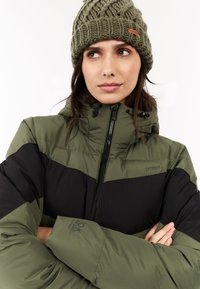 Vrouw met een olijfgroene gebreide muts en een bijpassende winterjas met twee tinten groen en zwart, met rits, met gekruiste armen en kijkt opzij.