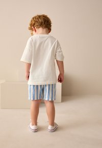 Camicia bianca a maniche corte, pantaloncini a righe blu e bianche e scarpe in tela bianche. Lo sfondo presenta un ambiente neutro con illuminazione soffusa.