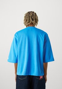 T-shirt de algodão azul com mangas curtas, corte descontraído e decote redondo. Detalhe de etiqueta notável na bainha. Cor sólida com uma textura suave.