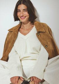 Brown suede jacket, cream knit sweater with a V-neck, and light-colored pants. Smooth texture, relaxed fit, and button accents on the jacket.