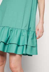 Robe verte sans manches avec des volants superposés à l'ourlet, confectionnée en tissu doux, avec une coupe décontractée et une texture lisse.
