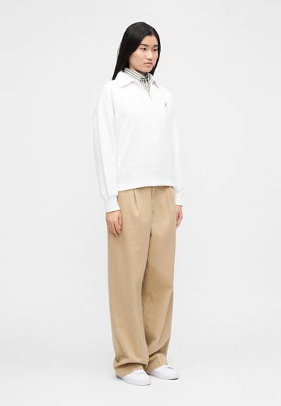 Sudadera blanca con cremallera y cuello, usada sobre una camisa de rayas. Pantalones de pierna ancha beige y zapatillas blancas completan el conjunto.