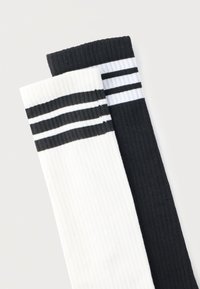 Ζευγάρι ριμπ κάλτσες τύπου φούτερ, μία λευκή με τρεις μαύρες ρίγες και μία μαύρη με τρεις λευκές ρίγες, τοποθετημένες επίπεδα σε ανοιχτό φόντο.