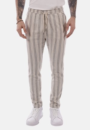 Uomo con pantaloni con coulisse beige e grigi a righe verticali e scarpe da ginnastica bianche, in piedi con le braccia rilassate lungo i fianchi.