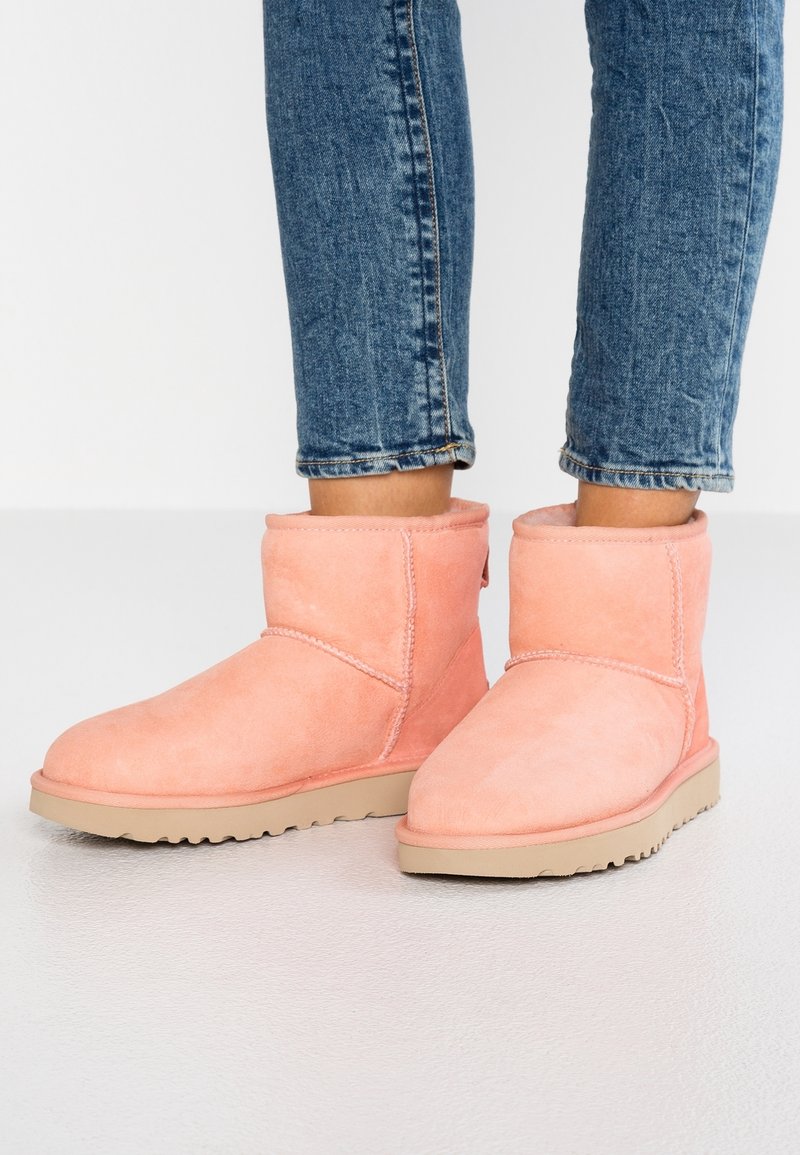Personne portant des bottines en daim rose pâle montant jusqu'à la cheville, avec des semelles beiges, et un jean bleu retroussé sur un sol gris clair.