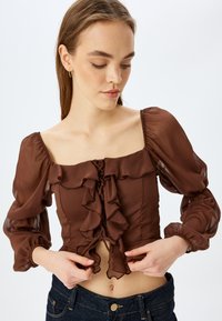 Braune, rüschige Bluse mit durchsichtigen Puffärmeln, quadratischem Ausschnitt und Schnürdetail vorne; getragen mit dunklen Jeans.