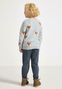 Kind met krullend blond haar kijkt weg, draagt een grijze trui met oranje vossengezichten, blauwe broek en bruine laarzen op een effen achtergrond.