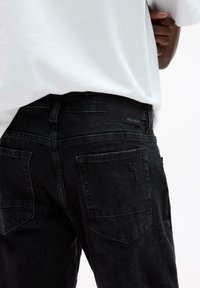 Person iført sorte denimjeans med baglommer og en hvid skjorte, hånd placeret på hoften, synligt logo på taljen.