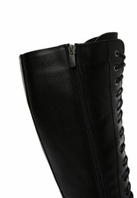 Stivale in pelle nera fino al ginocchio con lacci frontali e zip laterale per una facile vestibilità. Texture liscia con occhielli in metallo e dettagli di cucitura.