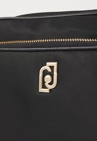 Schwarze Nylon-Tasche mit einem goldenen Reißverschluss und einem strukturierten goldenen Emblem auf der Vorderseite. Das Design ist elegant und minimalistisch.