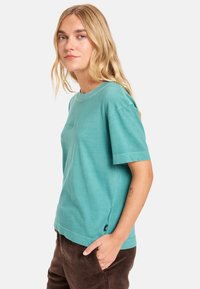 T-shirt corto di colore turchese in tessuto morbido con scollatura a girocollo e vestibilità rilassata, caratterizzato da un piccolo logo davanti e un'etichetta laterale.