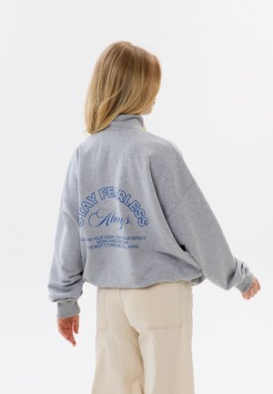 Kobieta o blond włosach stoi tyłem, ubrana w szarą bluzę z niebieskim napisem "STAY FEARLESS Always" i beżowe spodnie.