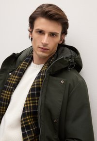 Parka verde con cappuccio, con chiusura frontale con cerniera. Sotto, un maglione bianco lavorato a trama e una sciarpa a quadri nera e gialla.