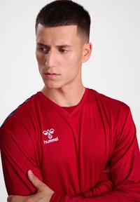 Rød langærmet atletisk skjorte med V-halsdesign, der har et hvidt Hummel-logo på brystet og kontrasterende sorte detaljer på ærmerne.