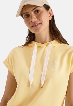 Jeune femme portant une casquette beige et un sweat à capuche jaune à manches courtes avec des cordons blancs et un texte circulaire sur la poitrine sur un fond uni.