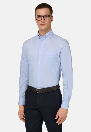 Mand iført sorte briller, lyseblå skjorte med knapper og lomme, sort bælte og mørke bukser, stående foran en ensfarvet baggrund.