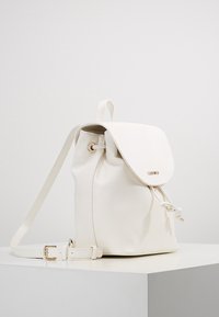 Vit faux läderryggsäck med lockstängning, dragsnodd och justerbar rem. Har guldfärgad hårdvara och en slät yta.