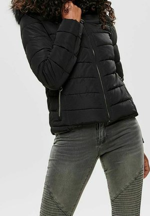 Personne portant une doudoune noire avec une capuche en fourrure et un jean gris avec des détails cousus sur les cuisses, se tenant devant un fond uni.