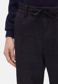 Homme portant un pantalon sombre à carreaux avec une taille élastique et un cordon de serrage, associé à une chemise à manches longues sombre, main reposant le long du corps.