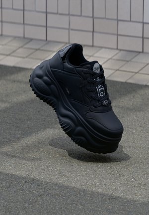 Czarny masywny sneaker na platformie z teksturowaną podeszwą i markowymi sznurówkami, umieszczony na tle ściany z płytek i asfaltowej nawierzchni.