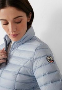 Chaqueta acolchada azul claro con costuras horizontales, cuello alto y cierre de cremallera. Cuenta con un parche de logo circular en la manga izquierda.