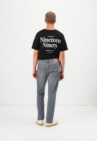 Hombre de pie mirando hacia atrás, vistiendo una camiseta negra con la frase blanca "Nineteen Ninety" y unos jeans grises con zapatillas blancas, sobre un fondo blanco.