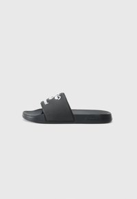 Čierne šľapky vyrobené z gumy s širokým remienkom, na ktorom je biele logo, plochá podrážka a hladký povrch. Minimalistický dizajn.