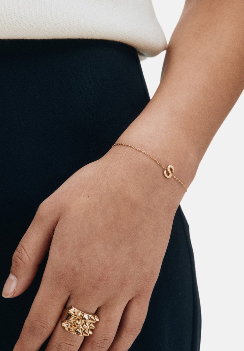 Bracciale in oro con ciondolo stilizzato a forma di 'S' accanto a un anello piramidale d'oro audace, posato su una mano contro uno sfondo nero.