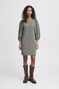 Abito a fantasia in verde tenue con motivi a diamante, maniche a sbuffo e taglio dritto, abbinato a stivali marroni fino al ginocchio.