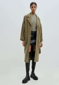 Olivgrüner oversized Mantel mit zwei фронт Taschen, getragen über einem ribbed hellgrünen Pullover und einem schwarzen Leder-Minirock. Schwarze knielange Stiefel.