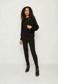 Μαύρη μπλούζα με μακριά μανίκια και λεπτομέρειες δαντέλας, συνδυασμένη με στενές μαύρες παντελόνες και μαύρες μποτάκια ως τον αστράγαλο. Το σύνολο διαθέτει απλό σχέδιο και υφή.