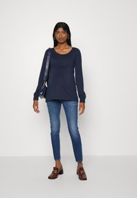 T-shirt azul-marinho de manga longa com decote arredondado e detalhe franzido, combinado com jeans skinny azuis e mocassins castanhos de salto. Bolsa preta.