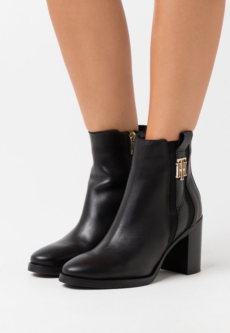 Femme portant des bottines en cuir noir à talons blocs avec un détail de logo doré sur le côté extérieur, se tenant contre un fond blanc.